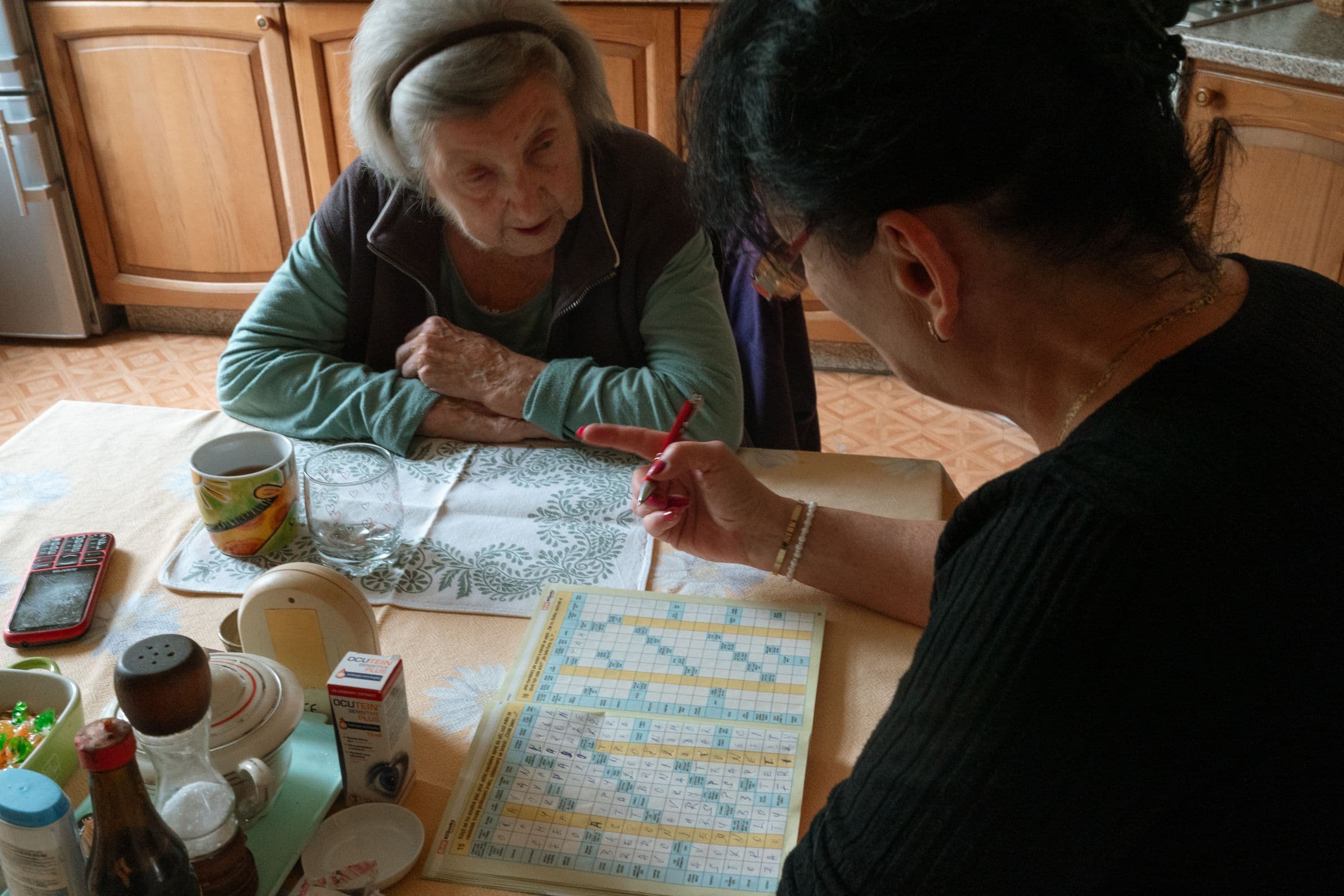 An older woman working on a crossword puzzle at her kitchen table with her caregiver.